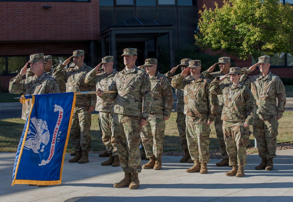 A group of United States Army soldiers in OCP (Operational Camouflage Pattern) uniforms are standing in formation during a military ceremony. Most soldiers are rendering a salute while one soldier holds the official United States Army flag (blue with gold fringe). The ceremony appears to be taking place outdoors on a paved area in front of a brick building.