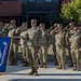 A group of United States Army soldiers in OCP (Operational Camouflage Pattern) uniforms are standing in formation during a military ceremony. Most soldiers are rendering a salute while one soldier holds the official United States Army flag (blue with gold fringe). The ceremony appears to be taking place outdoors on a paved area in front of a brick building.