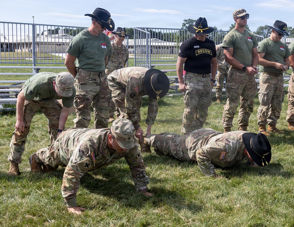 A group of U.S. Army soldiers are engaged in a physical training or assessment activity on a grass field. Two soldiers are performing push-ups on the ground while others stand over them observing or supervising. Several soldiers are wearing distinctive black cavalry-style cowboy hats, suggesting a cavalry unit affiliation.