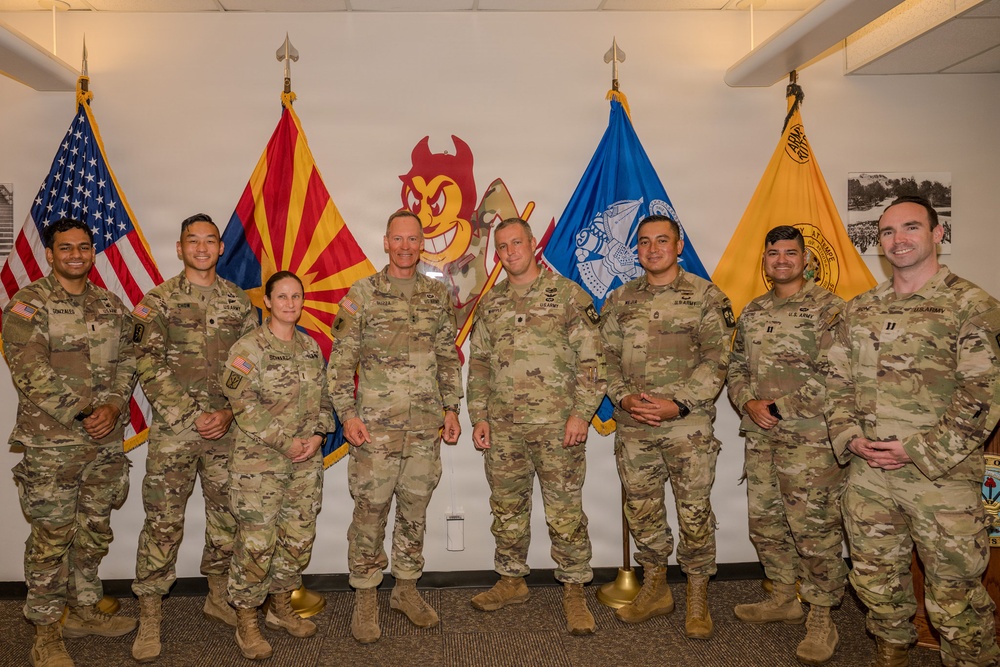 Eight U.S. Army soldiers in OCP (Operational Camouflage Pattern) uniforms pose for a formal group photo indoors. They are standing in front of multiple flags including the U.S. flag, Arizona state flag, a blue military unit flag, and a gold Army flag, with an Arizona State University Sun Devil mascot logo visible on the wall behind them. The setting appears to be an ROTC or military office at Arizona State University.
