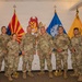 Eight U.S. Army soldiers in OCP (Operational Camouflage Pattern) uniforms pose for a formal group photo indoors. They are standing in front of multiple flags including the U.S. flag, Arizona state flag, a blue military unit flag, and a gold Army flag, with an Arizona State University Sun Devil mascot logo visible on the wall behind them. The setting appears to be an ROTC or military office at Arizona State University.
