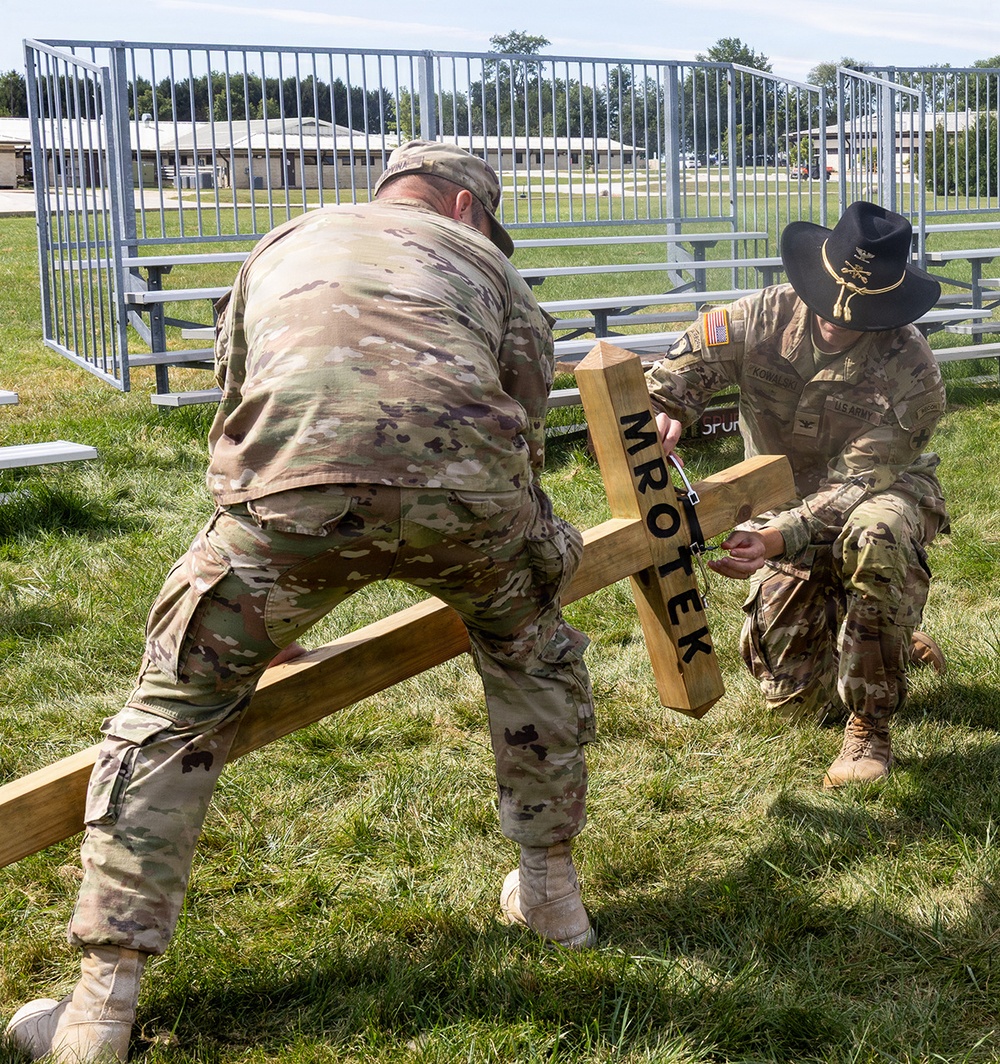 Two U.S. Army soldiers in OCP (Operational Camouflage Pattern) uniforms are working together to erect or position a wooden cross bearing the name 'MROTEK' on a grassy field. The soldier on the right wears a cavalry Stetson hat and a name tape reading 'KOWALSKI', and both wear U.S. Army insignia. The setting appears to be a military installation with bleachers and low buildings visible in the background.