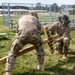 Two U.S. Army soldiers in OCP (Operational Camouflage Pattern) uniforms are working together to erect or position a wooden cross bearing the name 'MROTEK' on a grassy field. The soldier on the right wears a cavalry Stetson hat and a name tape reading 'KOWALSKI', and both wear U.S. Army insignia. The setting appears to be a military installation with bleachers and low buildings visible in the background.