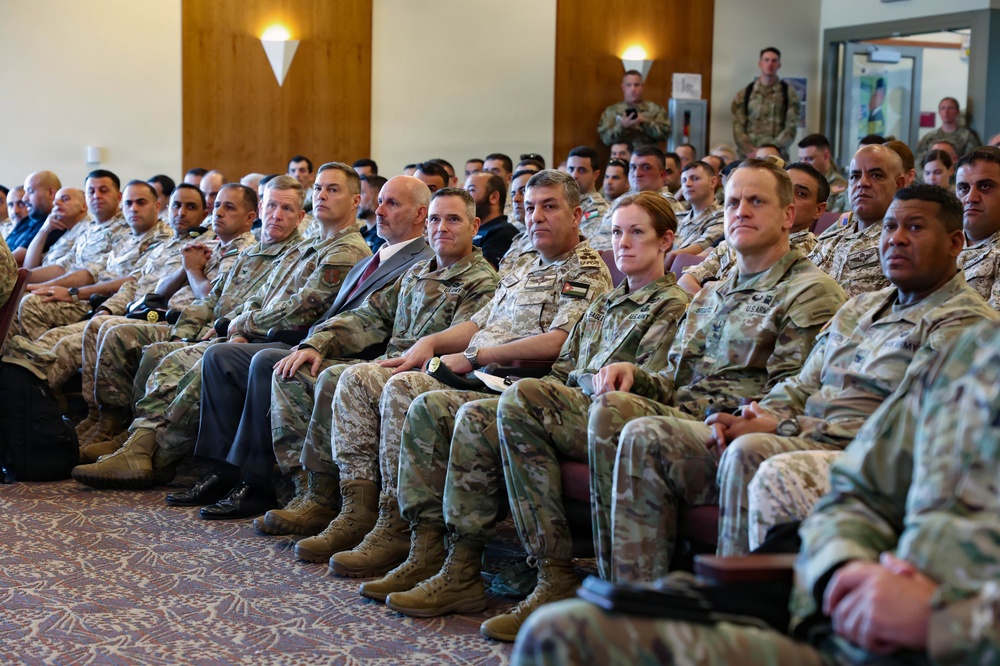 A large group of military personnel from what appears to be multiple nations are seated in rows in a conference or briefing room. The audience includes both men and women in various camouflage uniforms, with at least one civilian in a suit visible among them. The setting suggests a formal military briefing, ceremony, or multinational training event.