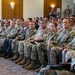 A large group of military personnel from what appears to be multiple nations are seated in rows in a conference or briefing room. The audience includes both men and women in various camouflage uniforms, with at least one civilian in a suit visible among them. The setting suggests a formal military briefing, ceremony, or multinational training event.