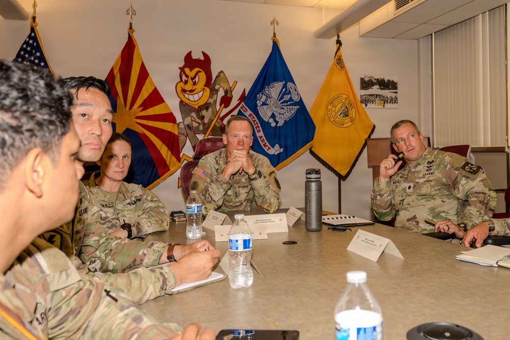 A group of U.S. Army personnel in camouflage uniforms are seated around a conference table in what appears to be a military briefing or meeting room. The setting includes multiple flags including the Arizona state flag, a United States Army flag, an Arizona State University flag (with the Sun Devil mascot visible), and an ASU unit flag. Name placards are visible on the table, suggesting a formal meeting or command briefing.