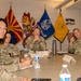 A group of U.S. Army personnel in camouflage uniforms are seated around a conference table in what appears to be a military briefing or meeting room. The setting includes multiple flags including the Arizona state flag, a United States Army flag, an Arizona State University flag (with the Sun Devil mascot visible), and an ASU unit flag. Name placards are visible on the table, suggesting a formal meeting or command briefing.