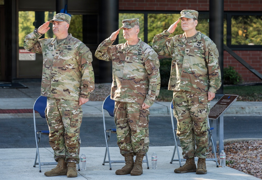 Three U.S. Army soldiers in Operational Camouflage Pattern (OCP) uniforms are standing at attention and rendering a salute during what appears to be a formal military ceremony. They are positioned in front of a brick building with blue folding chairs behind them and water bottles at their feet. A commemorative plaque or sign is visible to the right of the formation.