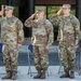 Three U.S. Army soldiers in Operational Camouflage Pattern (OCP) uniforms are standing at attention and rendering a salute during what appears to be a formal military ceremony. They are positioned in front of a brick building with blue folding chairs behind them and water bottles at their feet. A commemorative plaque or sign is visible to the right of the formation.