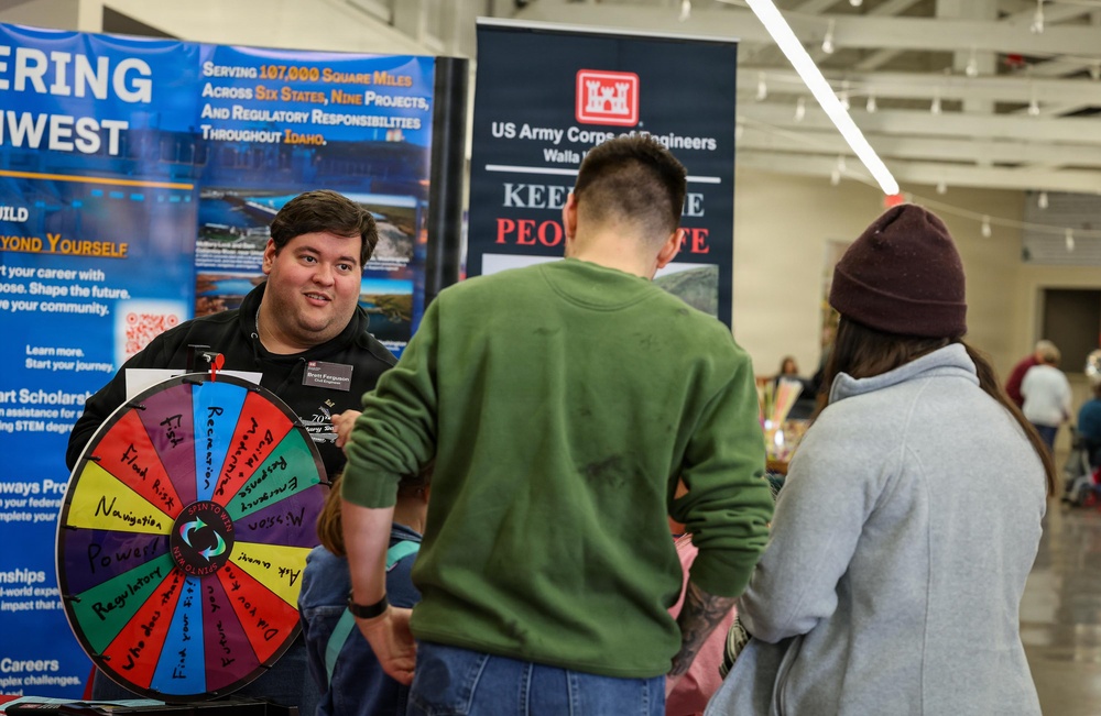 A recruiter or representative from the US Army Corps of Engineers (Walla Walla district) is engaging with two visitors at what appears to be a career fair or recruitment event. The representative is using a colorful prize/trivia spin wheel as an interactive engagement tool. Banners in the background reference engineering projects across Idaho and the Pacific Northwest.