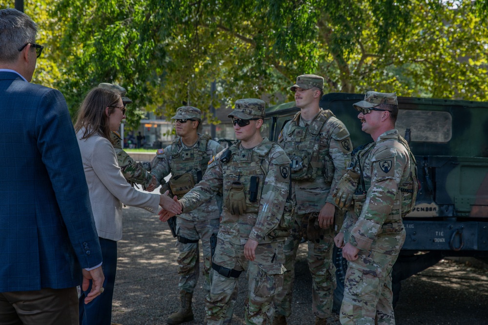 A civilian woman in a beige blazer is shaking hands with uniformed U.S. Army soldiers in OCP (Operational Camouflage Pattern) uniforms during what appears to be an official greeting or visit. A civilian man in a blue suit stands nearby observing the interaction. The setting is an outdoor area with trees and a military HMMWV (Humvee) vehicle visible in the background.