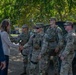 A civilian woman in a beige blazer is shaking hands with uniformed U.S. Army soldiers in OCP (Operational Camouflage Pattern) uniforms during what appears to be an official greeting or visit. A civilian man in a blue suit stands nearby observing the interaction. The setting is an outdoor area with trees and a military HMMWV (Humvee) vehicle visible in the background.