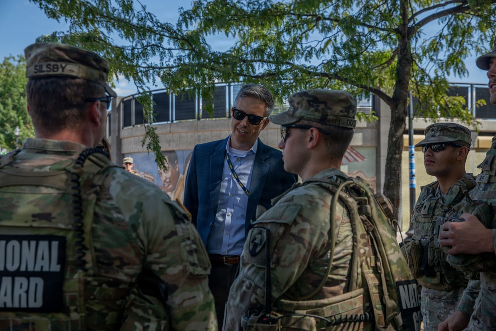 A civilian in a blue blazer and sunglasses is engaged in conversation with several National Guard soldiers in full combat uniforms and gear. The meeting appears to be taking place outdoors in an urban setting, possibly during a security or official visit. The civilian is wearing a lanyard/badge, suggesting an official or government role.