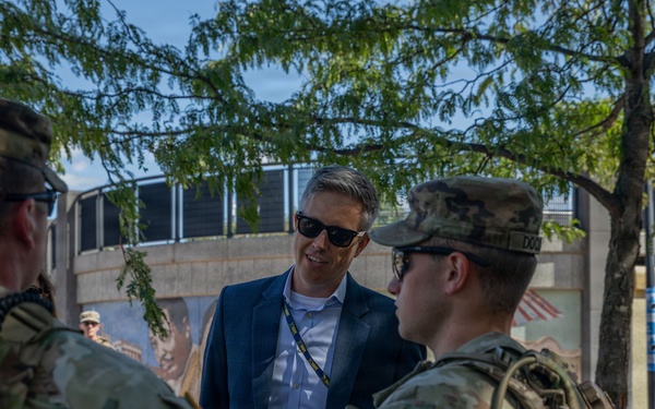 A civilian in a blue blazer and sunglasses is engaged in conversation with several National Guard soldiers in full combat uniforms and gear. The meeting appears to be taking place outdoors in an urban setting, possibly during a security or official visit. The civilian is wearing a lanyard/badge, suggesting an official or government role.