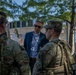 A civilian in a blue blazer and sunglasses is engaged in conversation with several National Guard soldiers in full combat uniforms and gear. The meeting appears to be taking place outdoors in an urban setting, possibly during a security or official visit. The civilian is wearing a lanyard/badge, suggesting an official or government role.