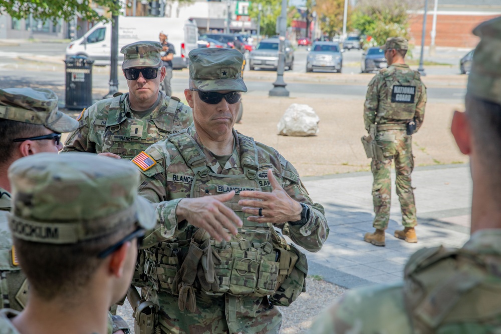 A group of U.S. Army National Guard soldiers are gathered on a city street, with a central figure wearing a name tape reading 'BLANCHARD' and displaying a general officer star on their cap, appearing to brief or address the group. A soldier in the background has 'NATIONAL GUARD' clearly visible on their vest. The setting appears to be an urban area with vehicle traffic visible in the background.
