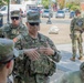 A group of U.S. Army National Guard soldiers are gathered on a city street, with a central figure wearing a name tape reading 'BLANCHARD' and displaying a general officer star on their cap, appearing to brief or address the group. A soldier in the background has 'NATIONAL GUARD' clearly visible on their vest. The setting appears to be an urban area with vehicle traffic visible in the background.