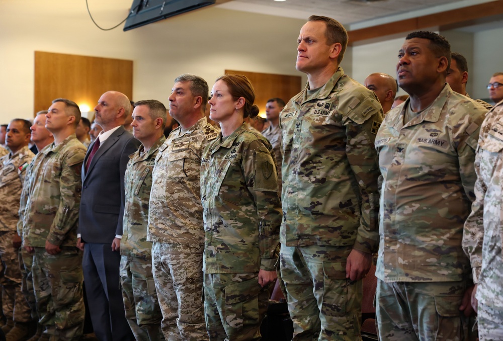 A group of military personnel, predominantly U.S. Army soldiers in OCP (Operational Camouflage Pattern) uniforms, are standing at attention in what appears to be an indoor ceremony or formal gathering. One individual in the front row is wearing a different digital camouflage pattern, suggesting a foreign military representative. A civilian in a suit is also visible among the group.