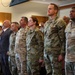 A group of military personnel, predominantly U.S. Army soldiers in OCP (Operational Camouflage Pattern) uniforms, are standing at attention in what appears to be an indoor ceremony or formal gathering. One individual in the front row is wearing a different digital camouflage pattern, suggesting a foreign military representative. A civilian in a suit is also visible among the group.