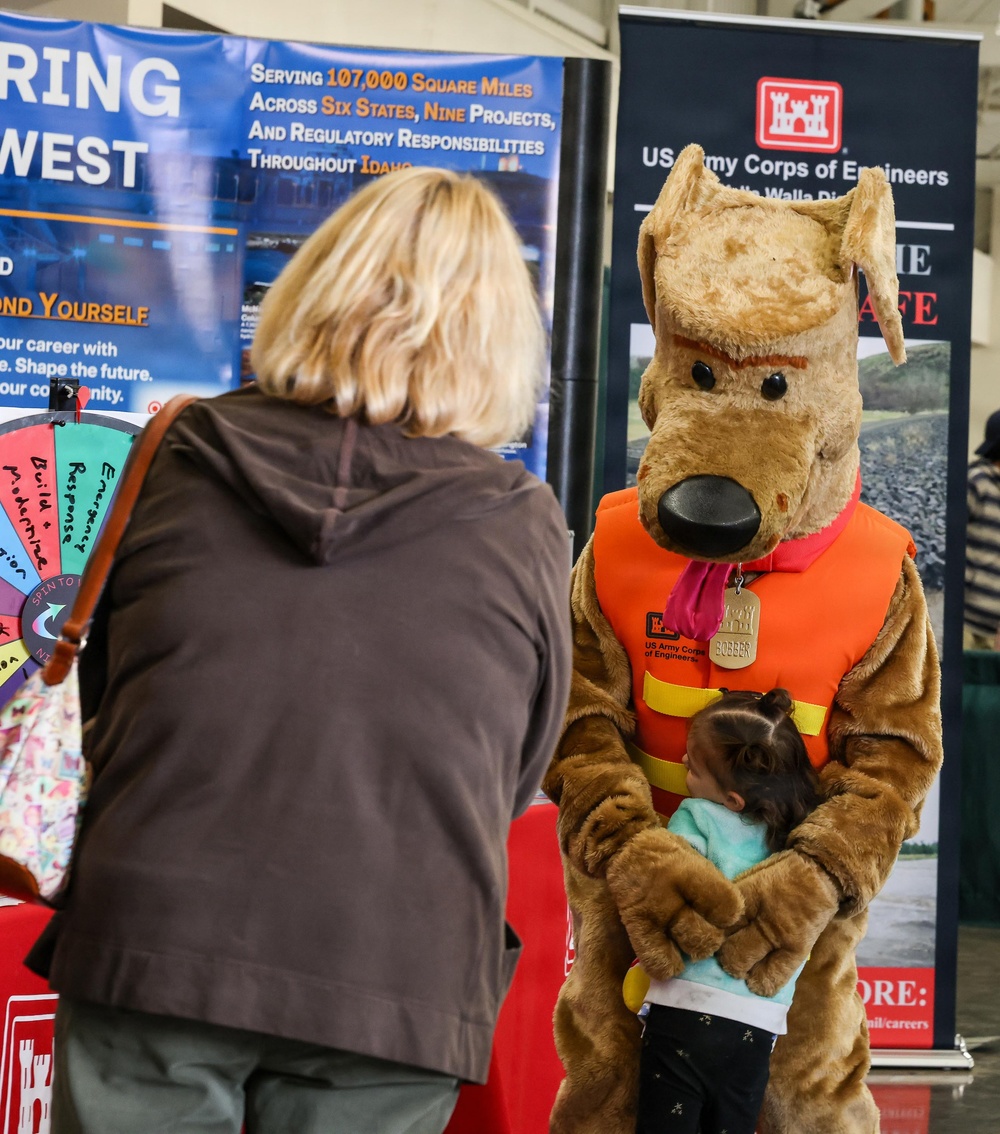 A US Army Corps of Engineers outreach event featuring their mascot 'Bobber,' a dog character wearing an orange life vest, hugging a small child. A blonde woman in a brown hoodie stands nearby at a booth with informational banners and a prize spin wheel. The event appears to be a public safety and recruitment fair promoting water safety and career opportunities.