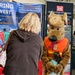 A US Army Corps of Engineers outreach event featuring their mascot 'Bobber,' a dog character wearing an orange life vest, hugging a small child. A blonde woman in a brown hoodie stands nearby at a booth with informational banners and a prize spin wheel. The event appears to be a public safety and recruitment fair promoting water safety and career opportunities.