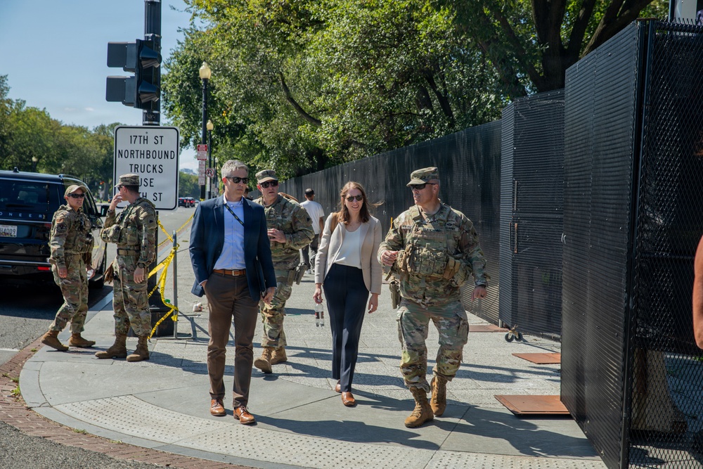 A group of military personnel in camouflage uniforms accompany two civilians — a man in a blue blazer and a woman in business casual attire — along a sidewalk near what appears to be a secured perimeter with black fencing. A street sign indicates the location is near 17th Street, suggesting a Washington D.C. setting. The scene suggests an official security or inspection walkthrough in a controlled area.