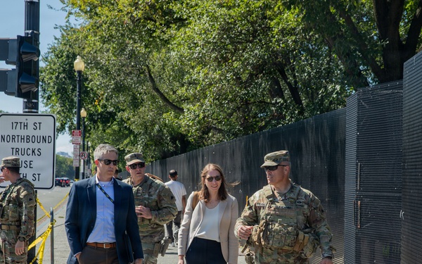 A group of military personnel in camouflage uniforms accompany two civilians — a man in a blue blazer and a woman in business casual attire — along a sidewalk near what appears to be a secured perimeter with black fencing. A street sign indicates the location is near 17th Street, suggesting a Washington D.C. setting. The scene suggests an official security or inspection walkthrough in a controlled area.