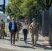 A group of military personnel in camouflage uniforms accompany two civilians — a man in a blue blazer and a woman in business casual attire — along a sidewalk near what appears to be a secured perimeter with black fencing. A street sign indicates the location is near 17th Street, suggesting a Washington D.C. setting. The scene suggests an official security or inspection walkthrough in a controlled area.