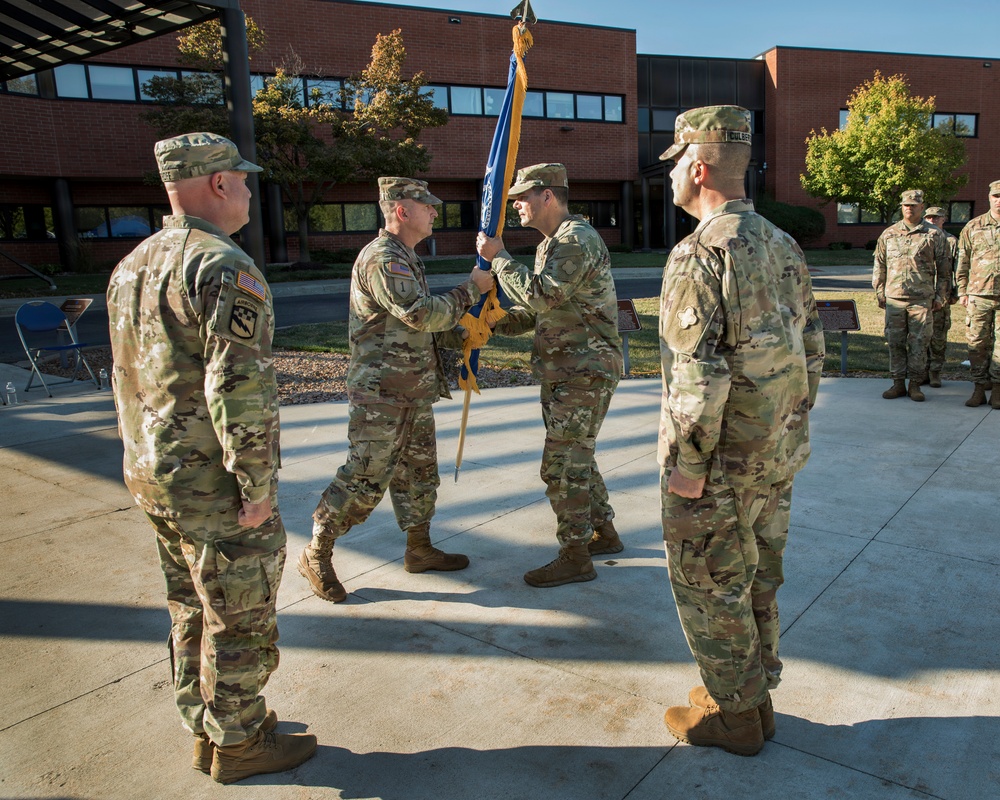 A military change of command ceremony is taking place outdoors in front of a brick building. Two soldiers are exchanging a blue and gold guidon (unit flag), which is the central symbolic act of a change of command. Additional soldiers stand in formation and as witnesses on both sides of the ceremony.
