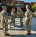 A military change of command ceremony is taking place outdoors in front of a brick building. Two soldiers are exchanging a blue and gold guidon (unit flag), which is the central symbolic act of a change of command. Additional soldiers stand in formation and as witnesses on both sides of the ceremony.