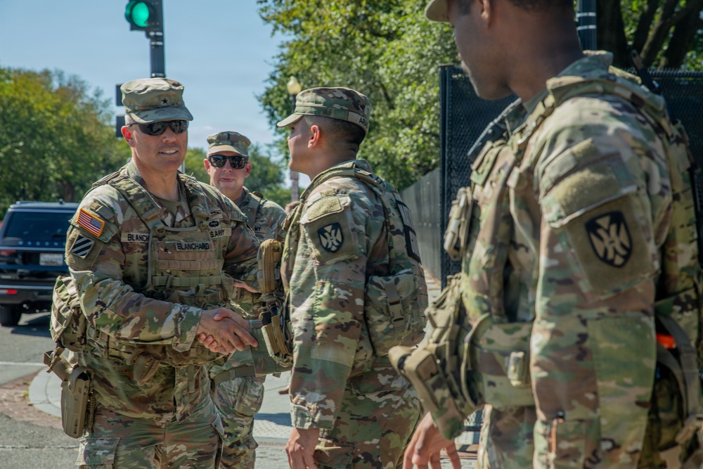 A group of U.S. Army soldiers in OCP (Operational Camouflage Pattern) uniforms are gathered on a city street. A senior officer identified by his name tape as 'BLANCHARD' is shaking hands with another soldier, suggesting a greeting or commendation. The setting appears to be an urban environment, possibly Washington D.C., based on the security fencing and surroundings.