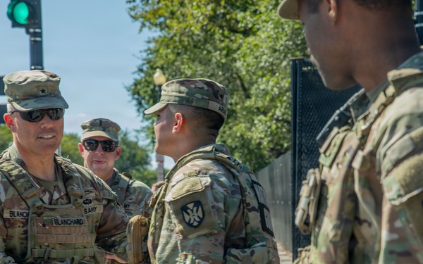 A group of U.S. Army soldiers in OCP (Operational Camouflage Pattern) uniforms are gathered on a city street. A senior officer identified by his name tape as 'BLANCHARD' is shaking hands with another soldier, suggesting a greeting or commendation. The setting appears to be an urban environment, possibly Washington D.C., based on the security fencing and surroundings.