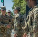 A group of U.S. Army soldiers in OCP (Operational Camouflage Pattern) uniforms are gathered on a city street. A senior officer identified by his name tape as 'BLANCHARD' is shaking hands with another soldier, suggesting a greeting or commendation. The setting appears to be an urban environment, possibly Washington D.C., based on the security fencing and surroundings.