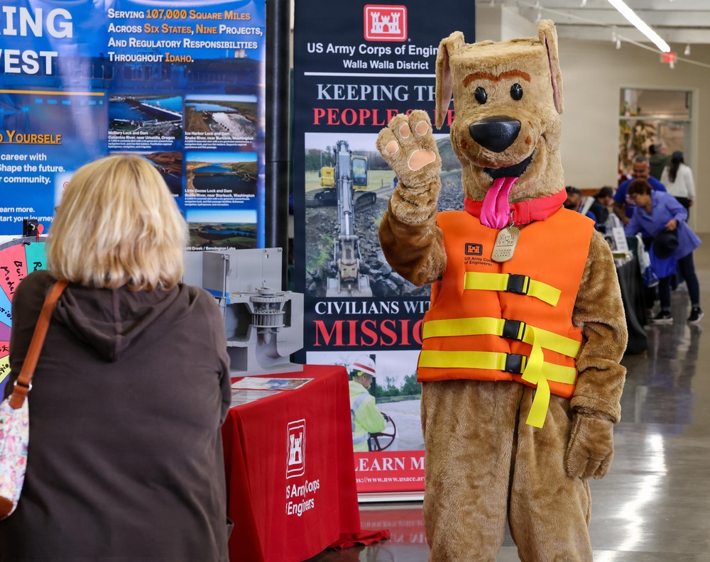 A US Army Corps of Engineers recruitment or outreach event featuring a dog mascot costume wearing an orange life vest with yellow straps. The mascot is waving at a woman with blonde hair who is facing away from the camera. The booth displays signage about the Walla Walla District and water safety messaging.