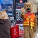 A US Army Corps of Engineers recruitment or outreach event featuring a dog mascot costume wearing an orange life vest with yellow straps. The mascot is waving at a woman with blonde hair who is facing away from the camera. The booth displays signage about the Walla Walla District and water safety messaging.