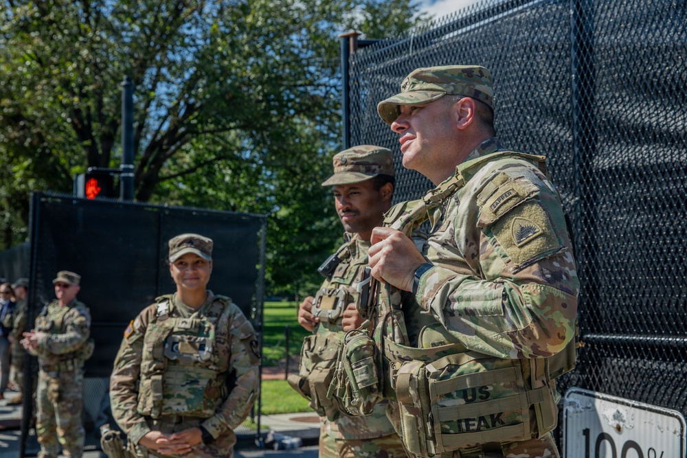 Three U.S. Army soldiers in OCP (Operational Camouflage Pattern) uniforms stand near a black security fence in an outdoor urban setting. The foreground soldier appears to be speaking or briefing, while others listen attentively. A red traffic signal and green trees are visible in the background.
