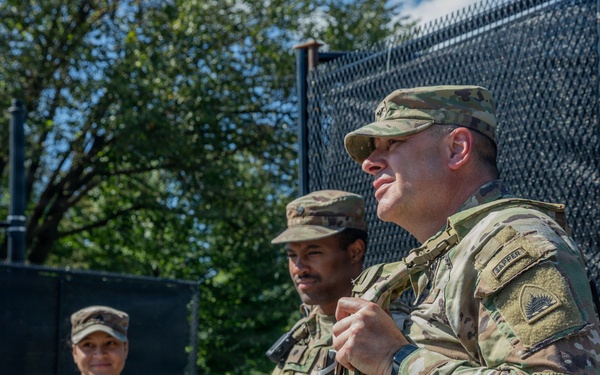 Three U.S. Army soldiers in OCP (Operational Camouflage Pattern) uniforms stand near a black security fence in an outdoor urban setting. The foreground soldier appears to be speaking or briefing, while others listen attentively. A red traffic signal and green trees are visible in the background.