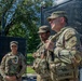 Three U.S. Army soldiers in OCP (Operational Camouflage Pattern) uniforms stand near a black security fence in an outdoor urban setting. The foreground soldier appears to be speaking or briefing, while others listen attentively. A red traffic signal and green trees are visible in the background.