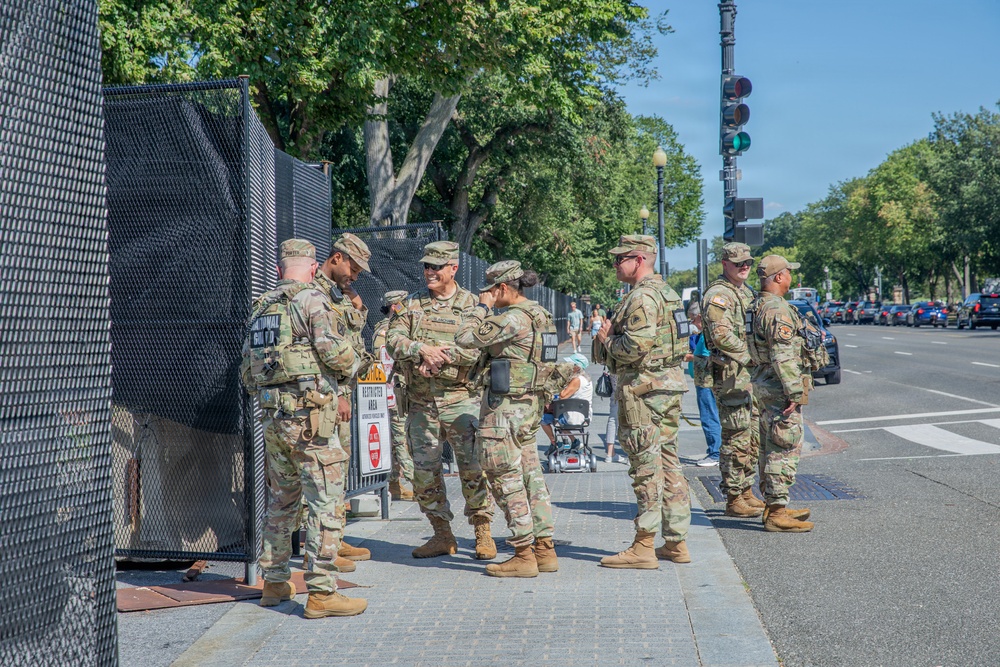 A group of approximately seven military personnel in camouflage uniforms are gathered on a sidewalk next to a security fence barrier. A sign reading 'Restricted Area' is visible near the fence, suggesting a secured perimeter in an urban setting. The scene appears to take place in Washington, D.C., based on the boulevard layout, tree-lined streets, and security infrastructure.