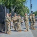 A group of approximately seven military personnel in camouflage uniforms are gathered on a sidewalk next to a security fence barrier. A sign reading 'Restricted Area' is visible near the fence, suggesting a secured perimeter in an urban setting. The scene appears to take place in Washington, D.C., based on the boulevard layout, tree-lined streets, and security infrastructure.