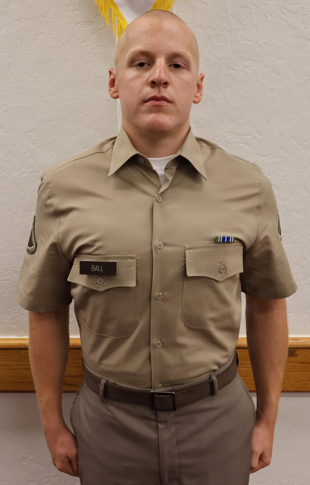 A young male in a khaki military-style uniform stands at attention in front of a white textured wall. He wears a name tag reading 'BALL' on his left chest pocket and a ribbon medal on his right chest. The photo appears to be a formal military or JROTC portrait.