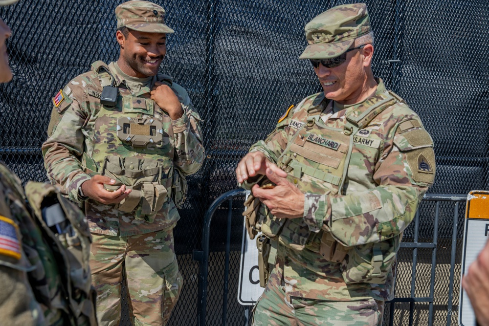 Two U.S. Army soldiers in MultiCam (OCP) uniforms are interacting near a security fence, appearing relaxed and engaged in conversation. The soldier on the right, identified by name tape as 'BLANCHARD' and wearing 'US ARMY' tape, appears to be a higher-ranking officer based on visible rank insignia on his cap. A third soldier is partially visible on the left edge of the frame.