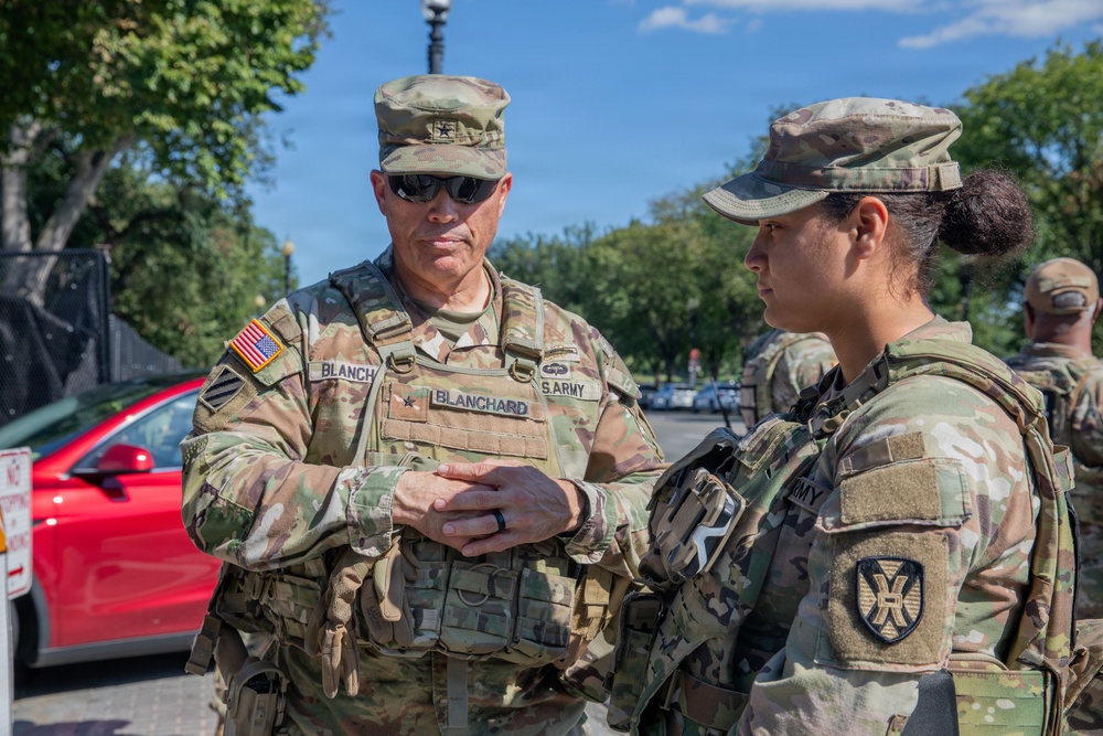Two U.S. Army soldiers in MultiCam (OCP) camouflage uniforms are engaged in conversation outdoors, likely at a security or ceremonial post. The soldier on the left displays the name tape 'BLANCHARD' and a single-star rank insignia indicating a Brigadier General. A third soldier is partially visible in the background, and the setting appears to be an urban area with trees and vehicles.