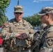 Two U.S. Army soldiers in MultiCam (OCP) camouflage uniforms are engaged in conversation outdoors, likely at a security or ceremonial post. The soldier on the left displays the name tape 'BLANCHARD' and a single-star rank insignia indicating a Brigadier General. A third soldier is partially visible in the background, and the setting appears to be an urban area with trees and vehicles.