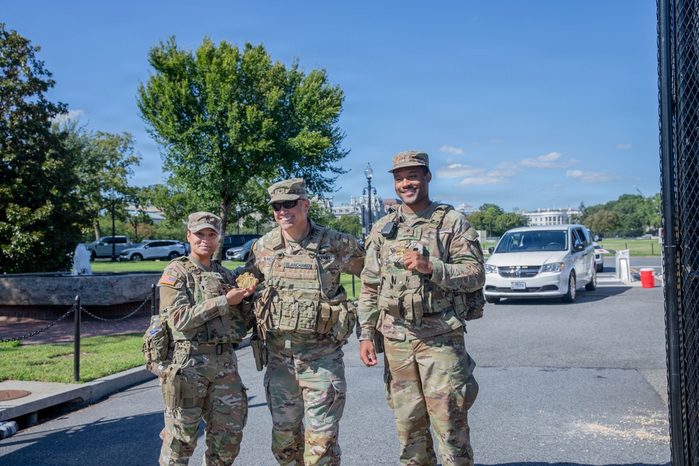 Three U.S. Army soldiers in OCP (Operational Camouflage Pattern) uniforms are posing together near a security fence, appearing to be on duty in Washington, D.C. They are smiling and holding what appear to be challenge coins or small items. The U.S. Capitol building and surrounding grounds are visible in the background.