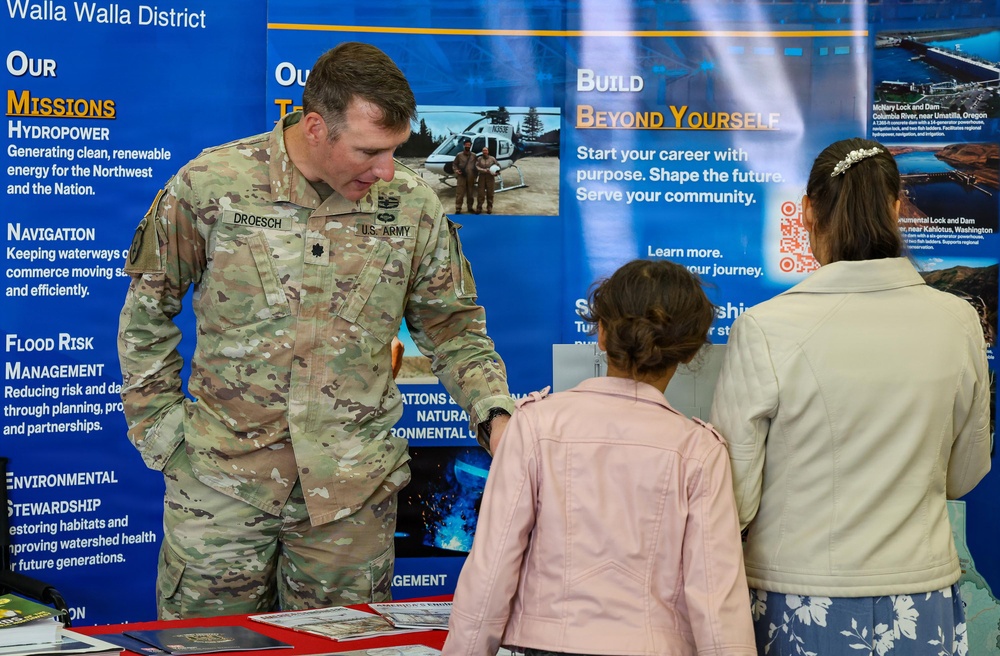A U.S. Army officer identified as 'Droesch' from the Walla Walla District is speaking with two civilian visitors at a recruitment or informational booth. The display features U.S. Army Corps of Engineers materials highlighting missions including hydropower, navigation, flood risk management, and environmental stewardship. The booth includes a 'Build Beyond Yourself' recruitment banner with a QR code.