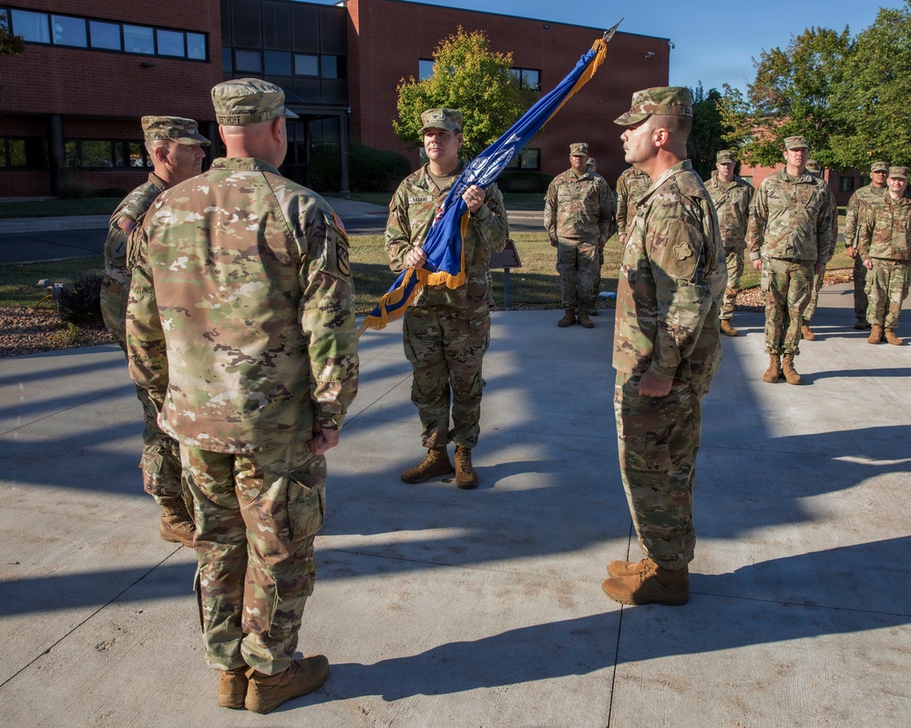 A military ceremony, likely a change of command or unit activation, is taking place outdoors on a paved area in front of a brick building. A soldier in the center is holding a blue unit guidon/flag with gold fringe, facing two senior personnel. Additional soldiers stand in formation in the background.
