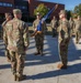 A military ceremony, likely a change of command or unit activation, is taking place outdoors on a paved area in front of a brick building. A soldier in the center is holding a blue unit guidon/flag with gold fringe, facing two senior personnel. Additional soldiers stand in formation in the background.