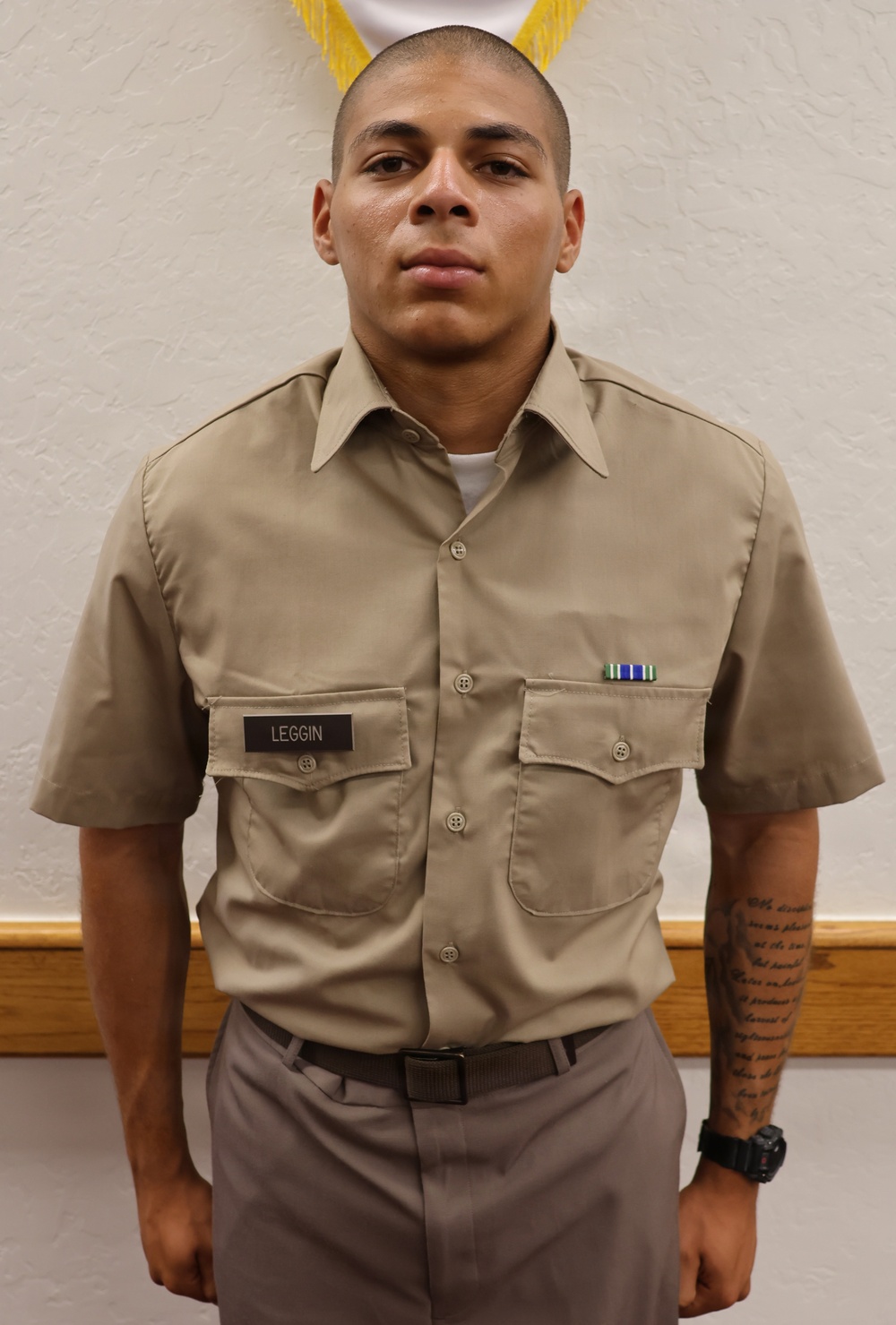 A young male service member in a khaki military uniform stands at attention in front of a white textured wall. He wears a name tag reading 'LEGGIN' on his left chest pocket and a small ribbon device on his right chest pocket. A tattoo is visible on his right forearm.