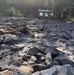 The image shows the aftermath of a debris flow or flash flood event in what appears to be a forested mountain area. Large boulders, uprooted trees, broken branches, and thick sediment/mud have been deposited across a wide area. A park or trailhead information kiosk with a green roof remains standing in the background, surrounded by the destruction.